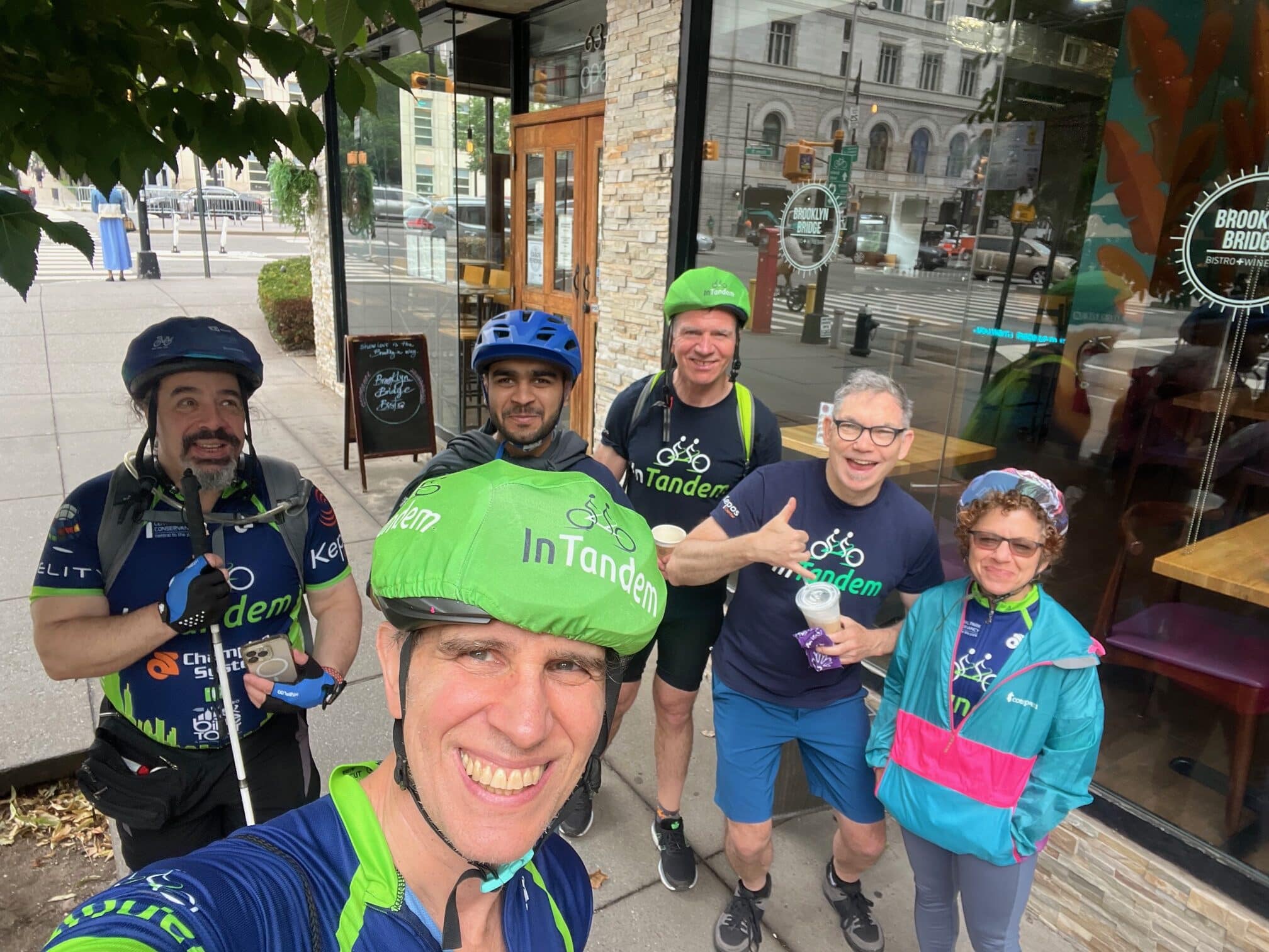 Six adults in cycling gear and helmets pose and smile for a group selfie on a sidewalk outside a storefront with large windows. One person holds a white cane. A city street and buildings are visible in the background.