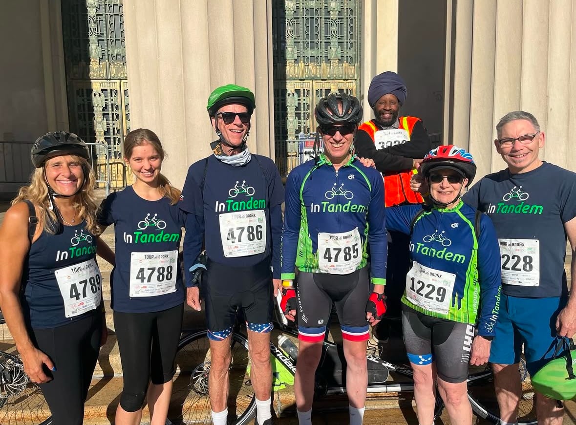 Seven people stand in front of a building, all wearing cycling gear and race numbers. Five wear matching In Tandem shirts. They are smiling and posing for the photo in bright sunlight.