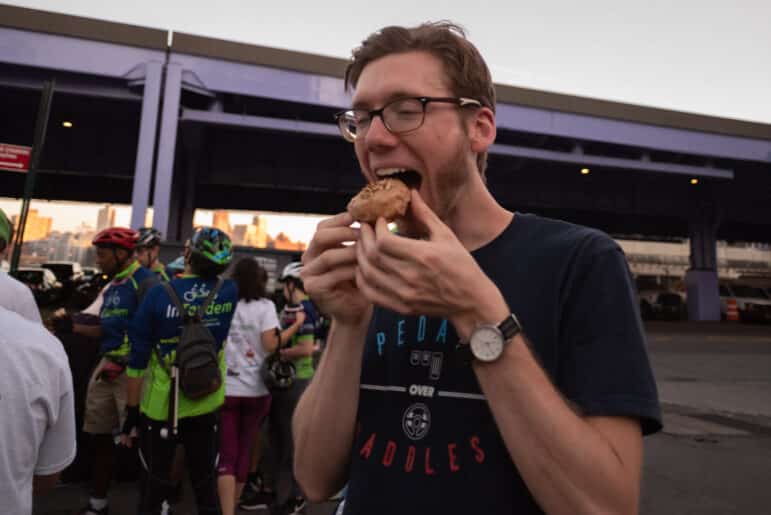 A man with glasses and a watch is eating a donut outdoors. In the background, several cyclists wearing helmets and colorful jerseys are gathered near bikes under a large overpass.