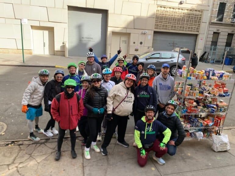 A group of people wearing helmets stand and pose for a photo on a city sidewalk next to a cart full of assorted canned goods and packaged food items. A few bicycles are visible, and buildings are in the background.