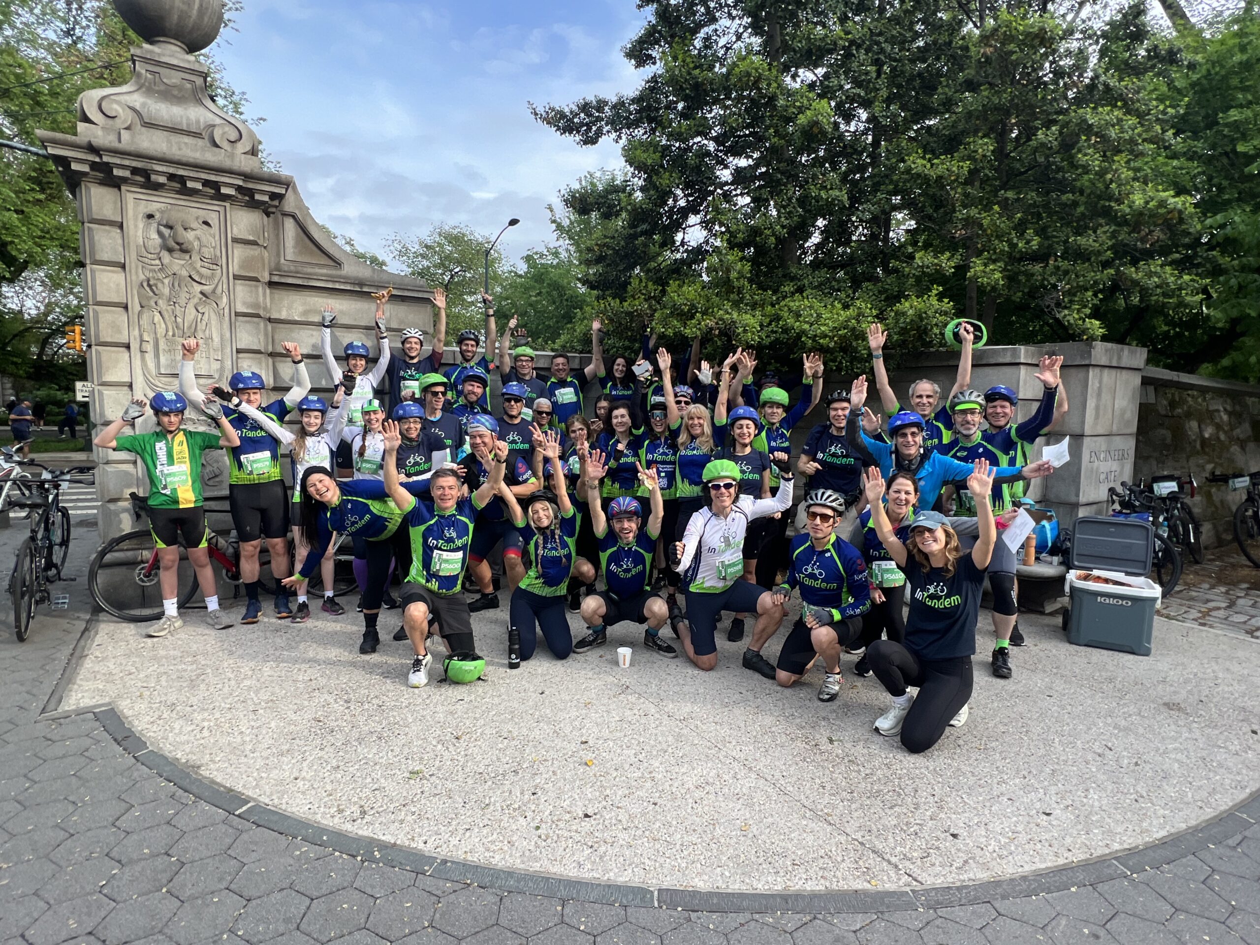 A large group of cyclists wearing helmets and sportswear pose together outdoors near a stone monument, many raising their arms and smiling, with bicycles and greenery in the background.