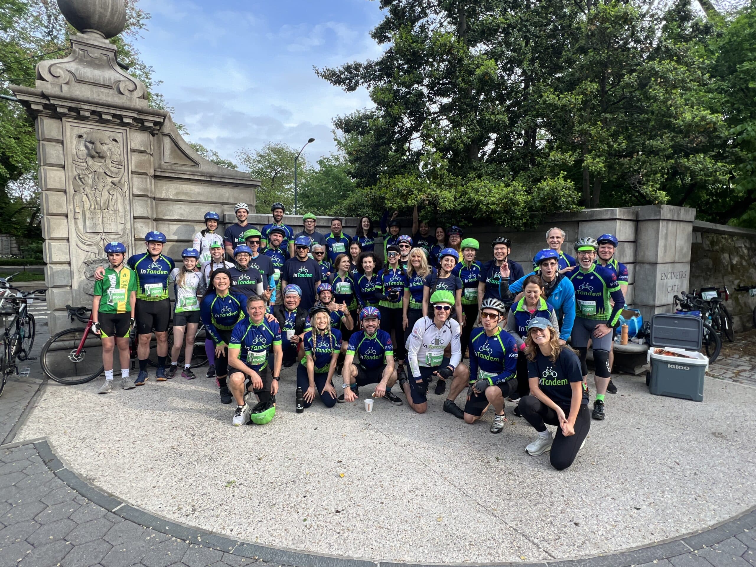 A large group of cyclists wearing matching blue and green jerseys and helmets pose together outdoors in front of a stone structure and greenery. Some have bikes, and a few people are kneeling in front.