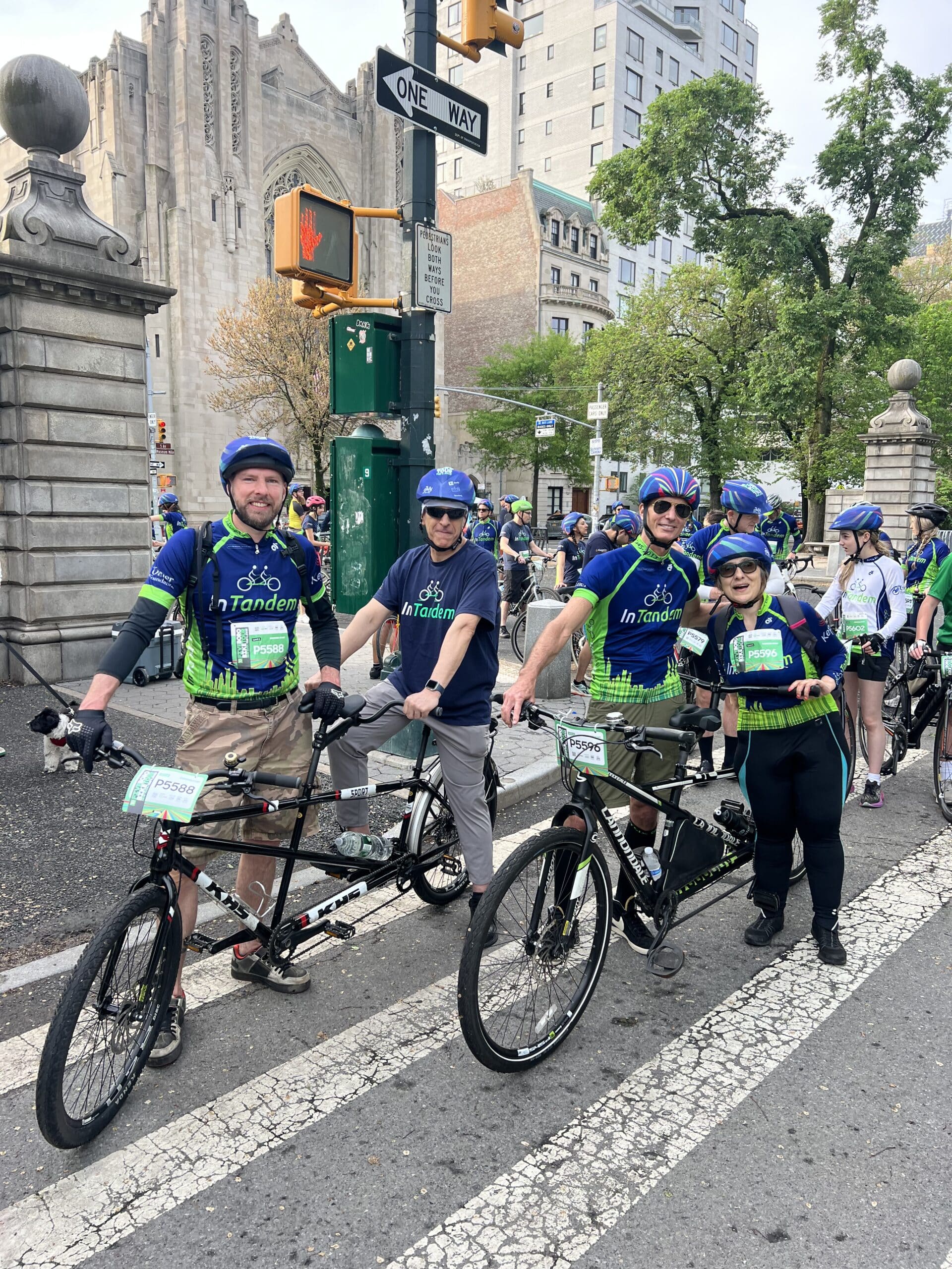 Three adults wearing bike helmets and InTandem shirts stand with tandem bicycles on a city street. Another woman stands beside them. Other cyclists are visible in the background near buildings and a traffic light.