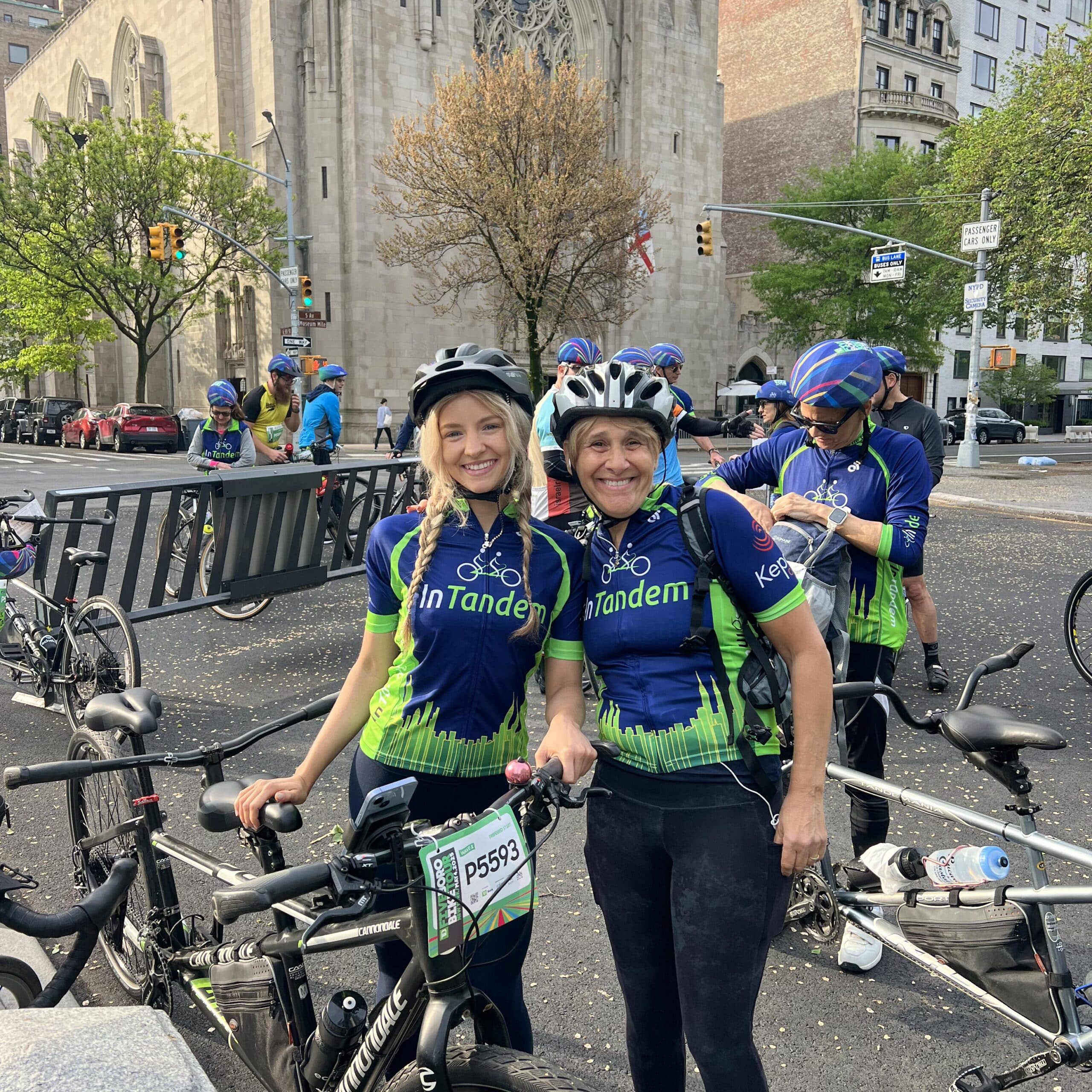 Two women in matching blue and green cycling jerseys and helmets stand with their bikes, smiling at the camera. Other cyclists are gathered behind them, and a large stone church is visible in the background.