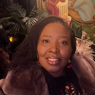 A woman with long braided hair smiles at the camera, wearing a fur coat and red earrings. There are green plants and a colorful painting in the background.
