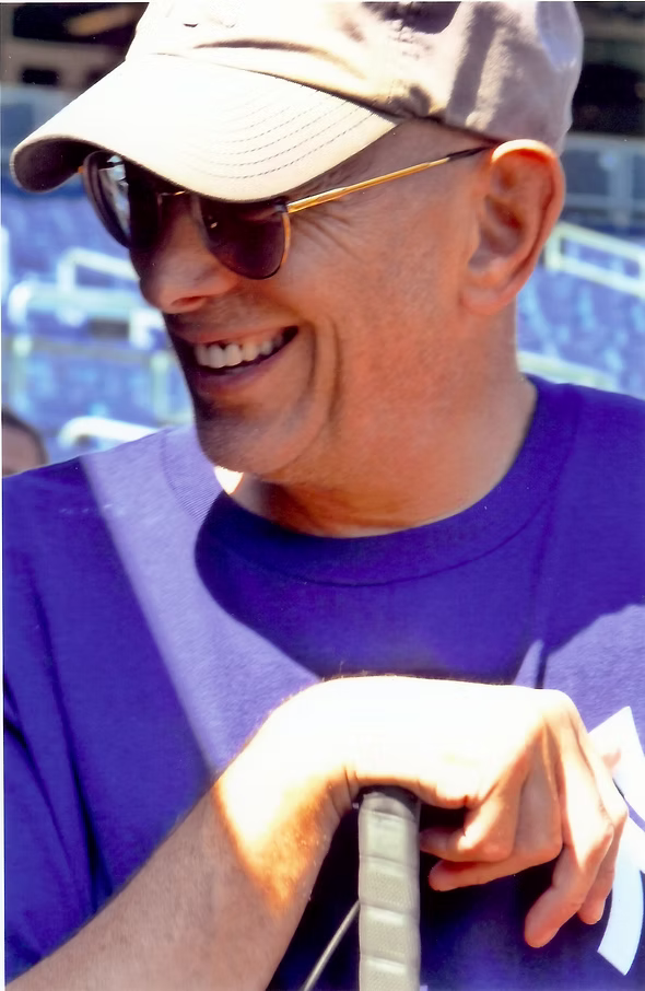 A person wearing a tan cap, sunglasses, and a purple T-shirt is smiling while leaning on a black object, with sunlight on their face and a blurred outdoor background.