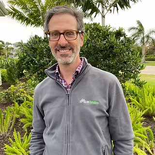 A man with glasses and gray hair stands outdoors in front of green bushes. He is wearing a gray zip-up jacket over a plaid shirt. Palm trees and a cloudy sky are visible in the background.