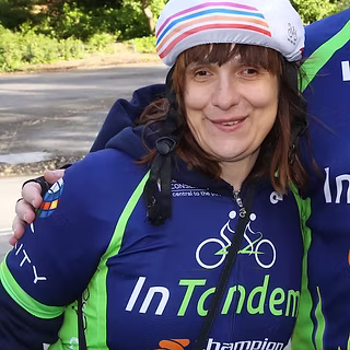 A woman wearing a white cycling cap and a blue InTandem cycling jersey stands outdoors, smiling. She is next to another person whose arm is around her shoulders. Trees and a paved area are in the background.