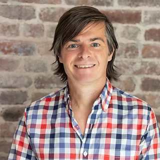 A person with medium-length brown hair is smiling at the camera, wearing a red, white, and blue plaid shirt, standing in front of a brick wall.
