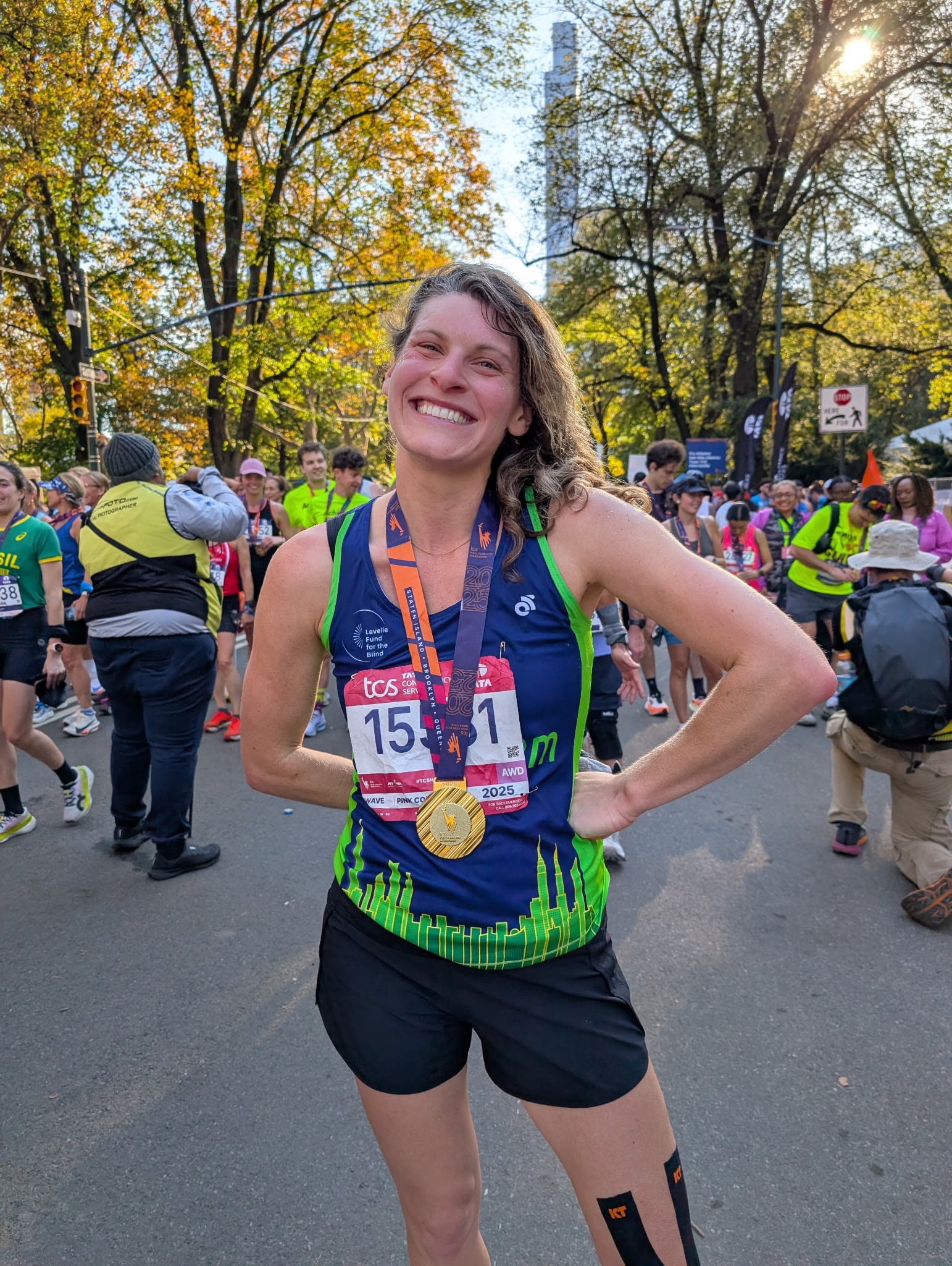 A woman in a blue and green running top, black shorts, and two medals smiles at the camera. She is standing in a street with other runners and trees in the background.