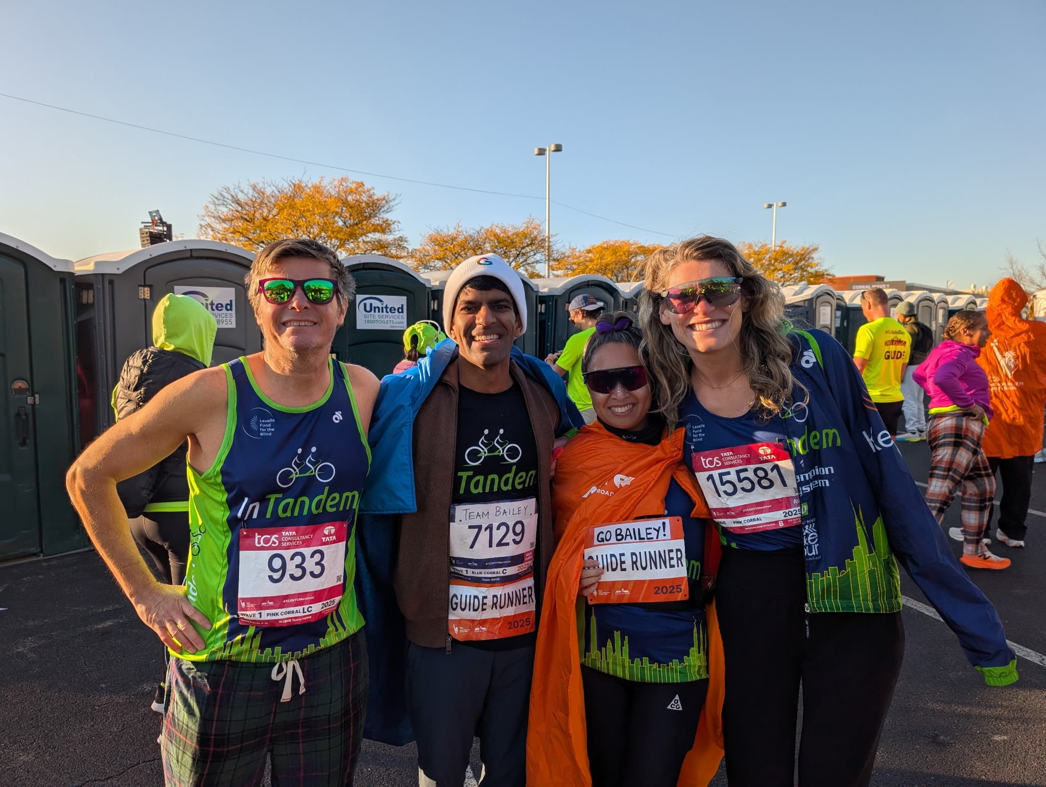 Four people wearing race bibs and “InTandem” shirts stand together smiling in front of portable toilets. Two wear “Guide Runner” vests. Other runners and volunteers are visible in the background.