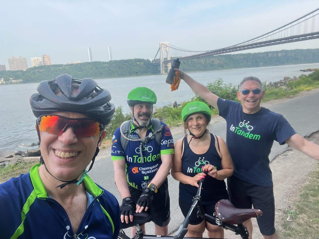 Four cyclists wearing InTandem shirts and helmets pose for a selfie by a river. A large bridge spans the background, and buildings and trees are visible across the water. One person smiles and holds up a cloth.