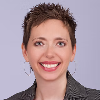 A person with short, spiky brown hair is smiling at the camera. They are wearing a gray blazer and large hoop earrings, with a light purple background behind them.