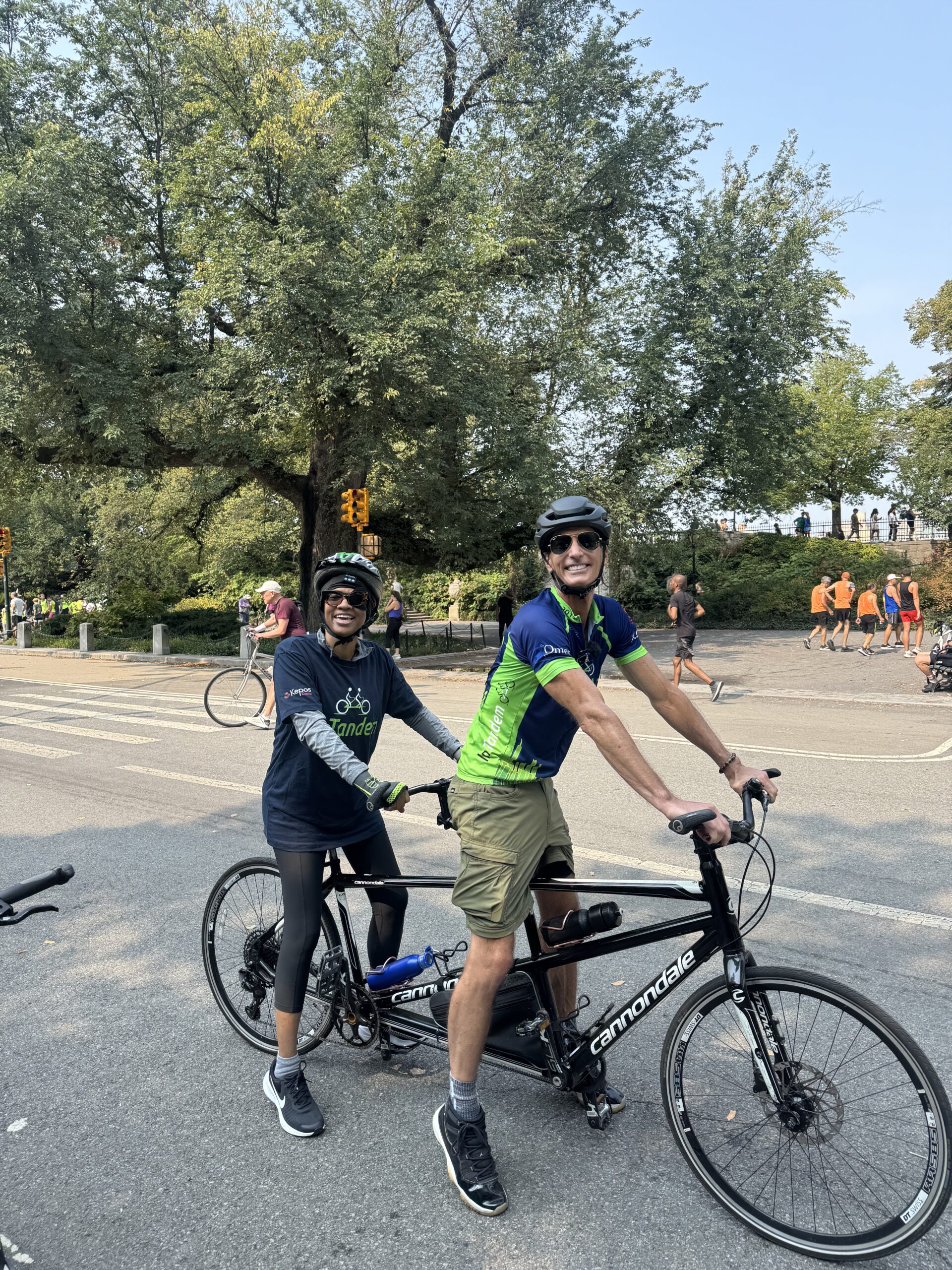 Two people wearing helmets and cycling gear are riding a tandem bicycle on a paved road. Trees and other cyclists are visible in the background on a sunny day.