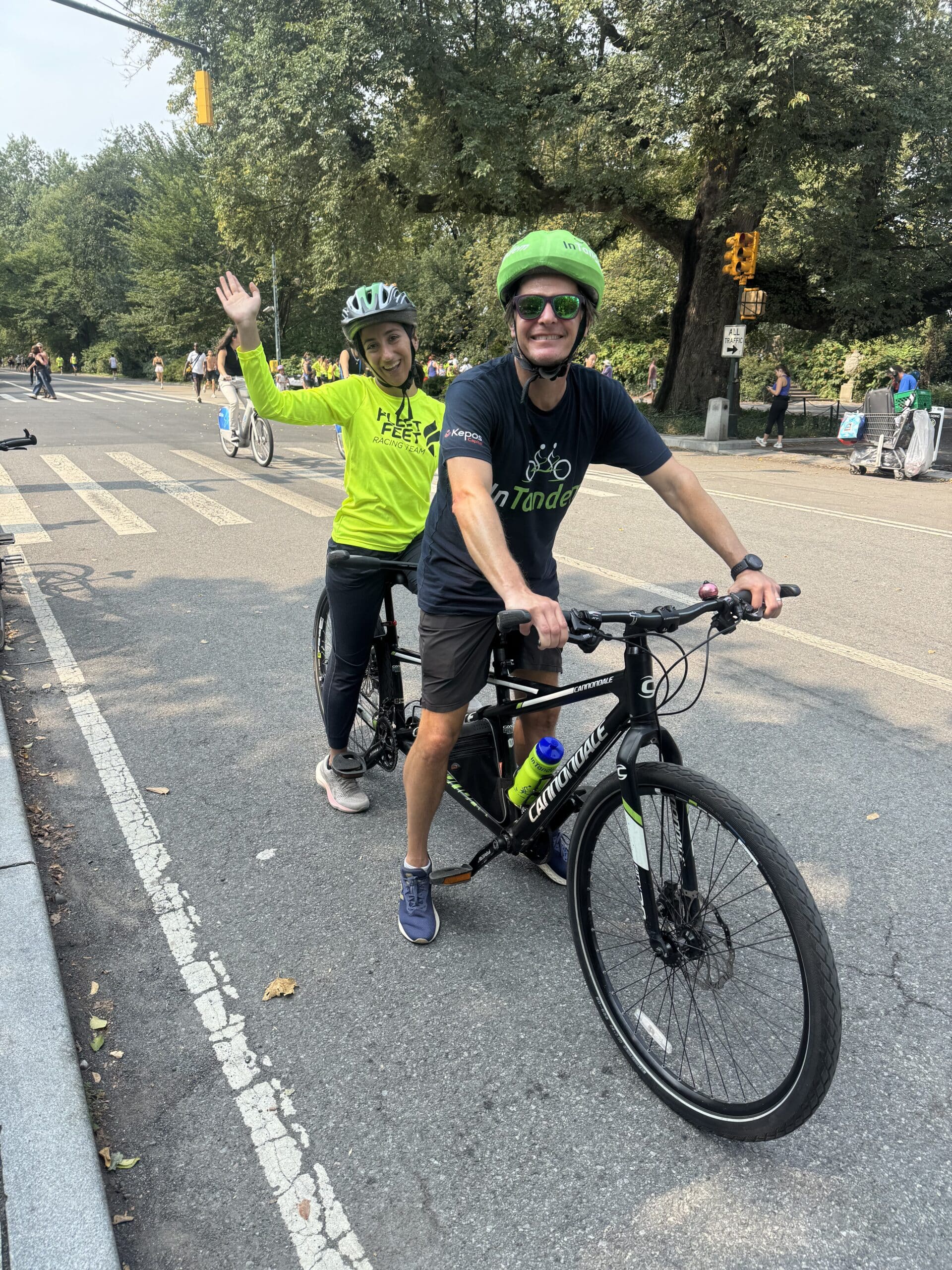 A smiling rider in an InTandem Cycling shirt steers a tandem bike while a participant in a green jersey waves to the camera on a Central Park road.