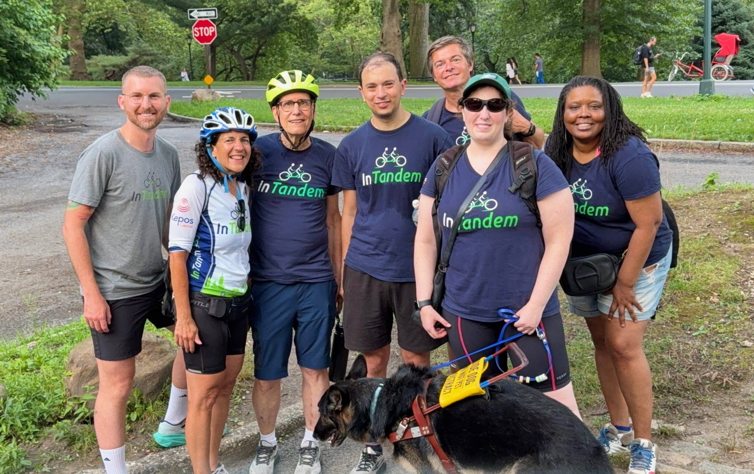 A group of seven people and a German Shepherd service dog stand in front of a park scene with a stop sign and trees. They are all wearing dark blue t-shirts with In Tandem logo.