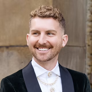 A person with short curly hair and a trimmed beard is smiling while wearing a dark velvet jacket, a white shirt, and decorative gold jewelry. The background appears to be a stone wall.
