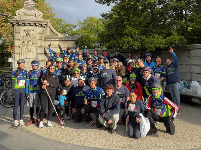 A large group of people wearing cycling gear pose together outdoors in front of a stone monument with greenery and trees in the background. Some are kneeling, and a few hold helmets or bikes.