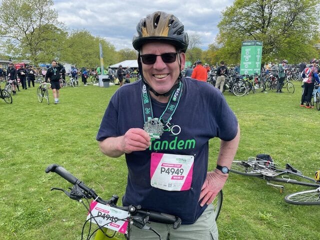 A person wearing a bike helmet and sunglasses stands outdoors holding a medal. They have a race number bib labeled P4949 and stand beside a bicycle, with other cyclists and bikes in the background.
