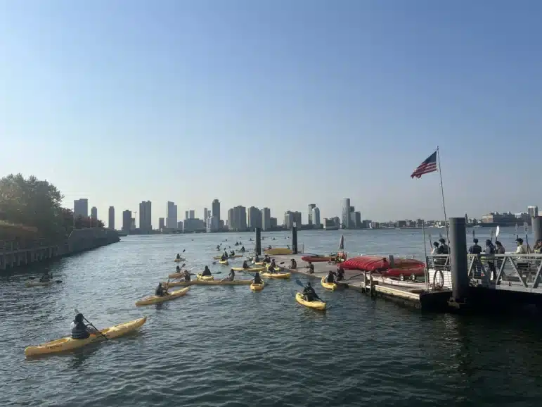People paddle yellow kayaks near a dock on a river, with a group of others gathered on the dock. An American flag flies nearby and a city skyline is visible in the distance under a clear blue sky.
