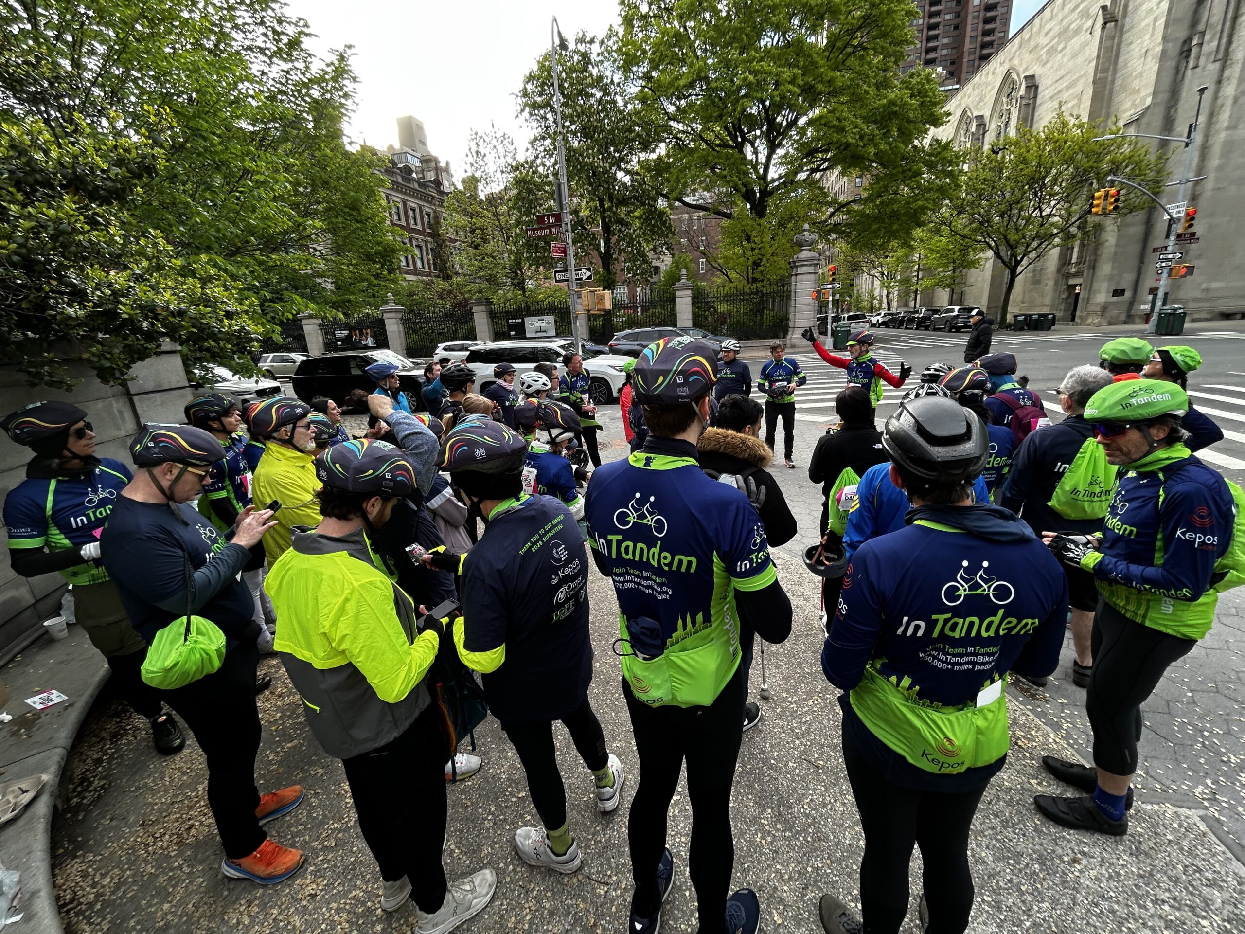 Four cyclists wearing InTandem shirts and helmets pose for a selfie by a river. A large bridge spans the background, and buildings and trees are visible across the water. One person smiles and holds up a cloth.