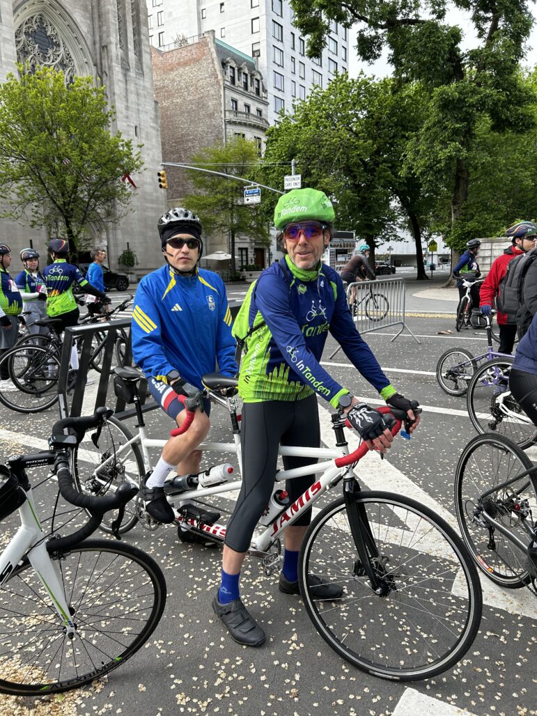Two cyclists stand on their tandem bike, both wearing blue and green jerseys and helmets.