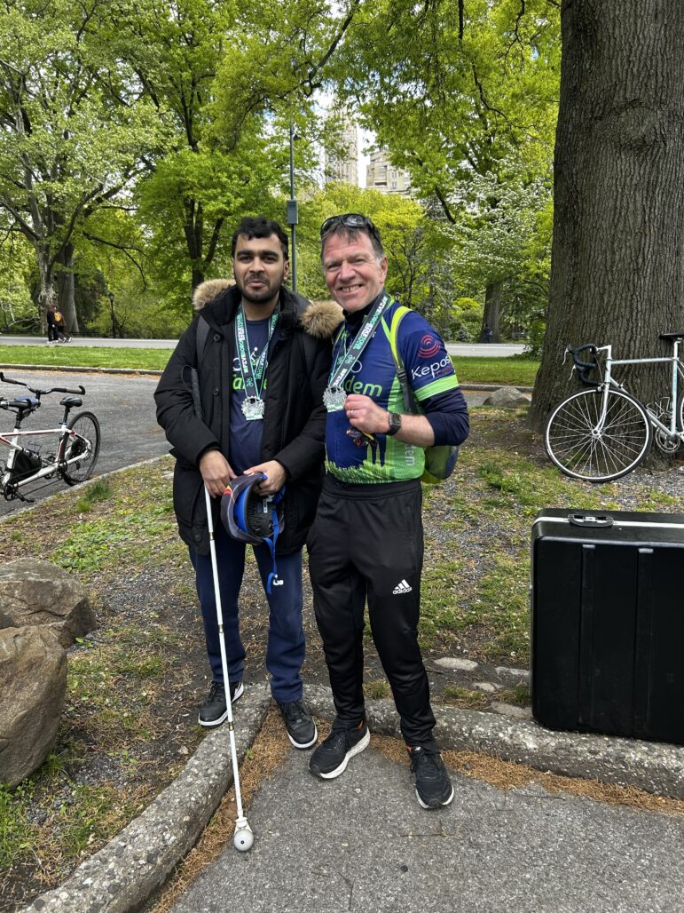 An InTandem Captain and Stoker stand together, posed for a picture, one holding their 5 Boro Medal up and smiling. The Stoker stands smiling holding his cane and wearing his medal.