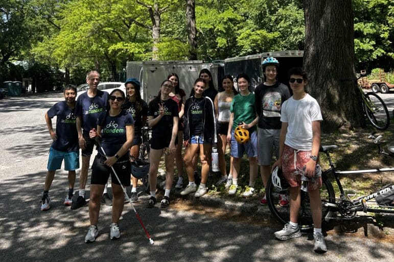 A group of twelve people, some holding bikes and wearing helmets, stand together outdoors on a sunny day near trees and a silver trailer. Most are smiling at the camera; some wear matching “tandem” shirts.