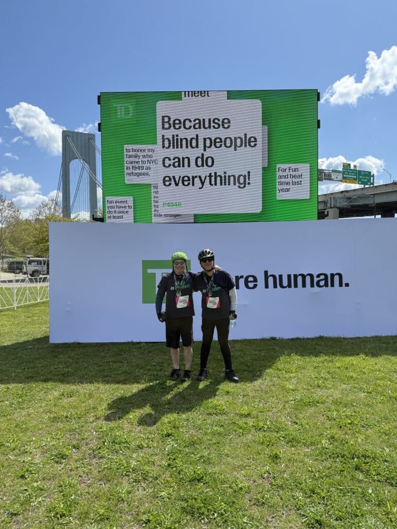Two cyclists pose in front of a large TD billboard that reads Because blind people can do everything! and a white sign with TD: Inspire human. The background includes a bridge, blue sky, and clouds.