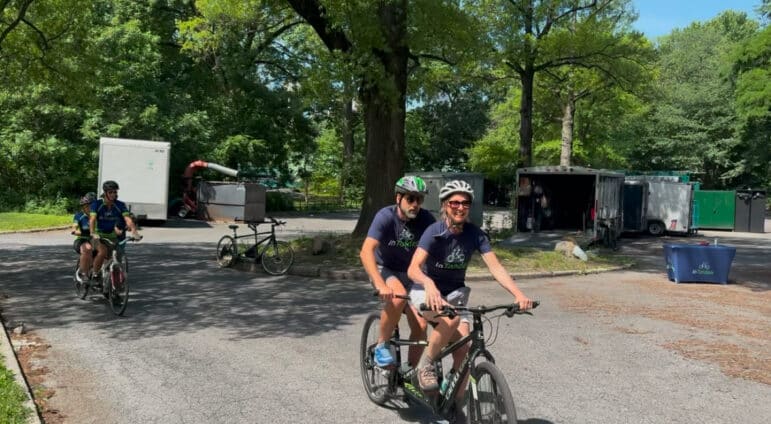 Two people wearing helmets and navy t-shirts ride a tandem bicycle on a paved path in a park. Trees and parked trailers are visible in the background. A single bike is parked to the left.
