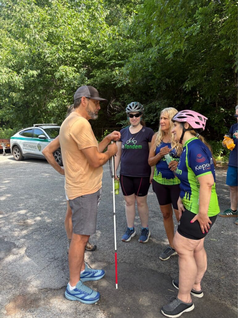 A man with a white cane stands and talks with four women in cycling gear in a parking area next to a NYC Parks car, with trees in the background.